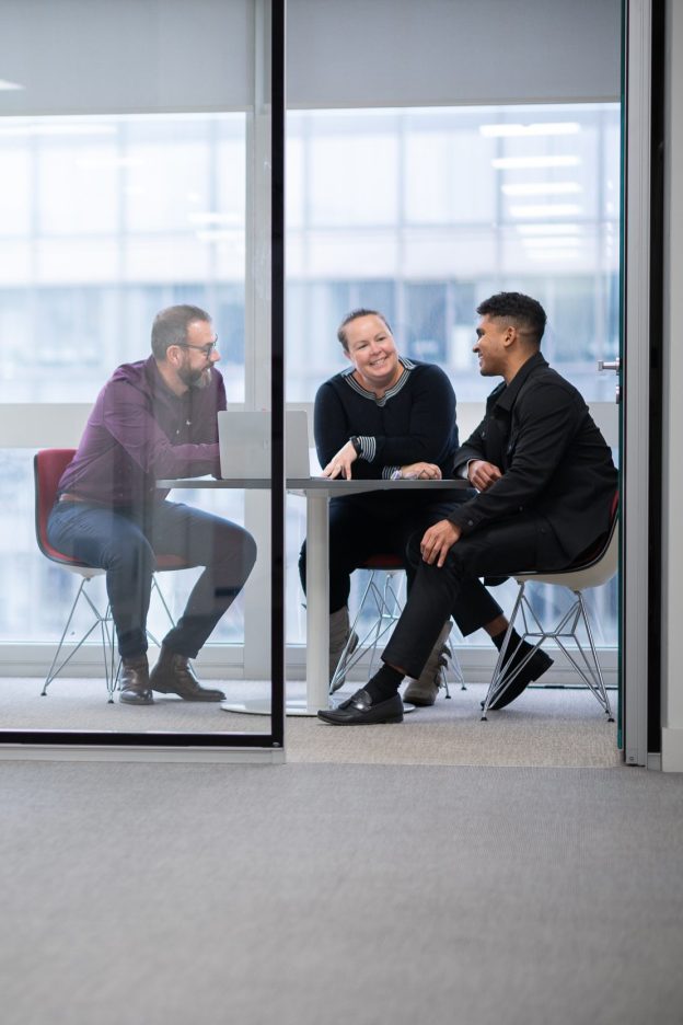 A candid shot from afar of three individuals sitting in a meeting behind glass doors